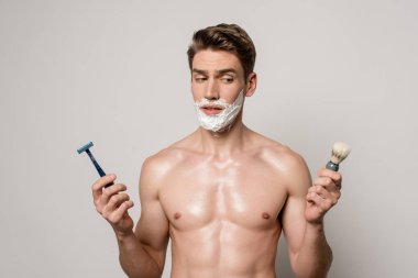 sexy man with muscular torso and shaving foam on face holding shaver and shaving brush isolated on grey clipart