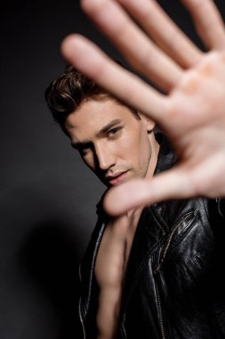 selective focus of sexy young man with muscular torso in biker jacket showing hand at camera on black background clipart