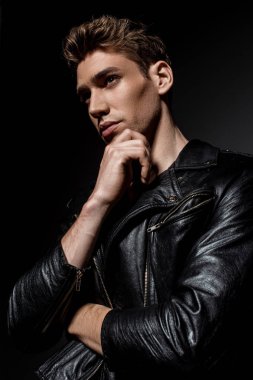 Motosiklet ceketli seksi genç adam çeneye dokunuyor ve siyah arka plana bakıyor.