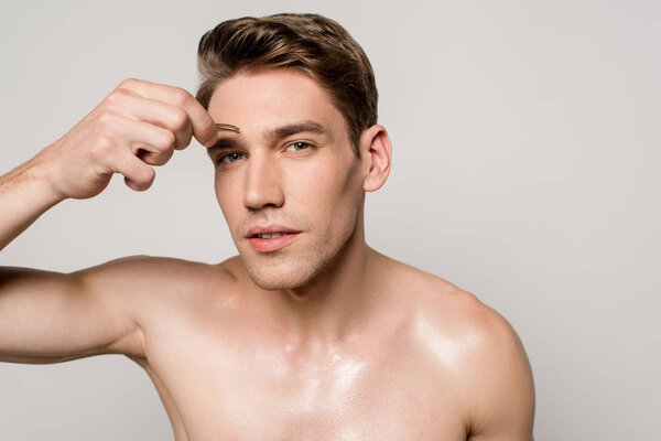 sexy man with bare torso pinching brows with tweezers isolated on grey