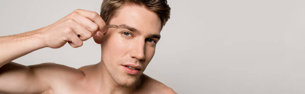 sexy man with bare torso pinching brows with tweezers isolated on grey, panoramic shot