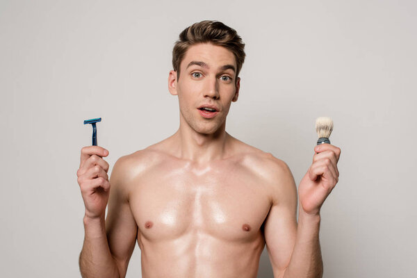 shocked sexy man with muscular torso holding shaver and shaving brush isolated on grey