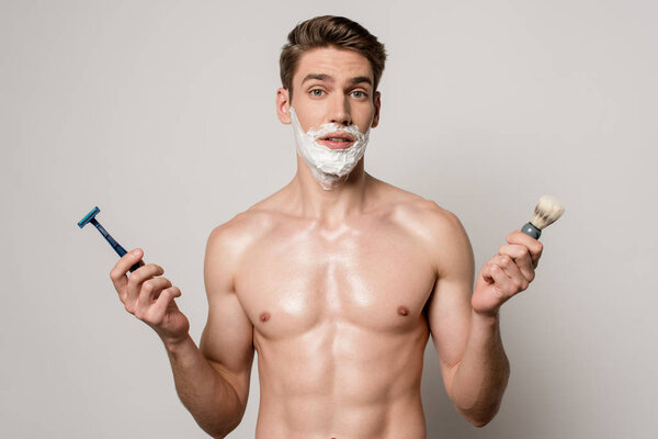 sexy man with muscular torso and shaving foam on face holding shaver and shaving brush isolated on grey