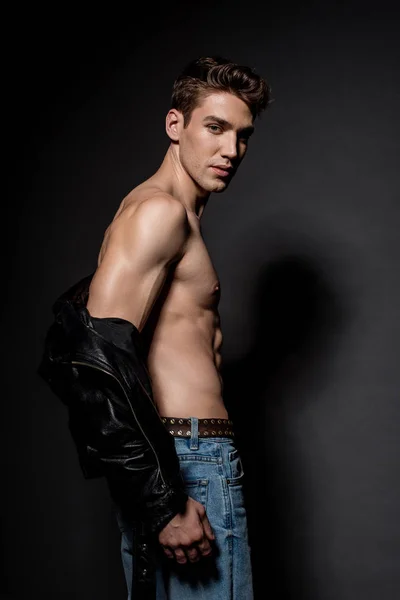 サイドビューのセクシーな若い男で筋肉質の胴体でバイカージャケットとジーンズで黒の背景 — ストック写真