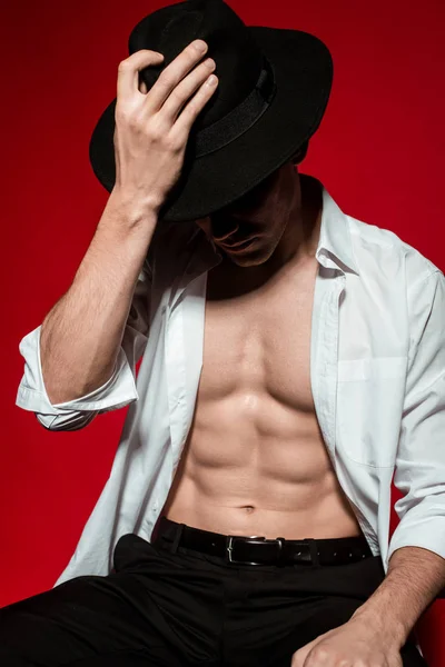 セクシーな若いですエレガントな男ですボタンなしシャツともに筋肉裸の胴着上の帽子の上に赤の背景 — ストック写真