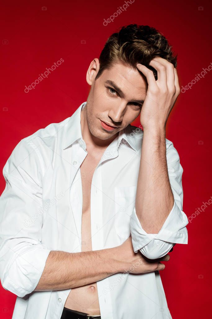 sexy young elegant man in unbuttoned shirt touching hair on red background