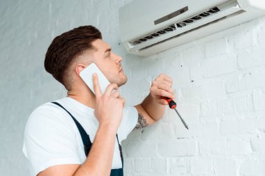 Akıllı telefonla konuşurken ve elinde tornavida tutarken klimaya bakan yakışıklı bir kurucu.