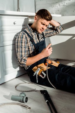 tired installer sitting on floor and holding metal hose near kitchen cabinet  clipart