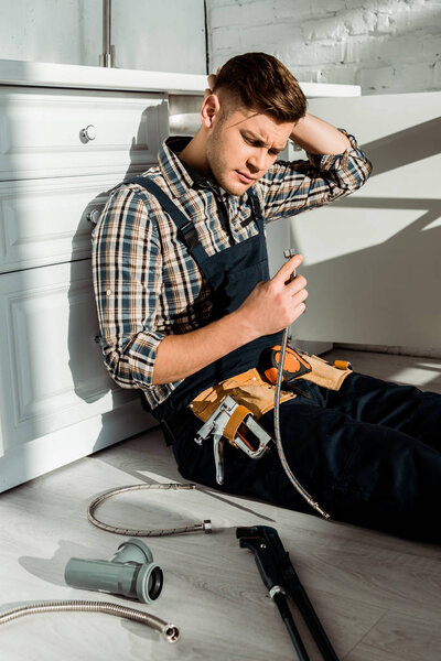 уставший монтажник сидит на полу и держит металлический шланг возле кухонного шкафа
 