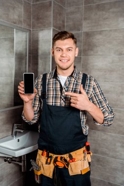 happy installer pointing with finger at smartphone with blank screen in bathroom  clipart