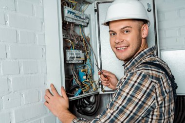 Kasklı mutlu elektrikçi elinde tornavidayla santralin yanında.  