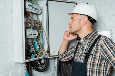 side view of pensive electrician touching face while looking at switchboard   clipart