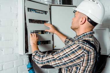 Güvenlik kasklı elektrikçi tuğla duvarın yanındaki santrale bakıyor. 