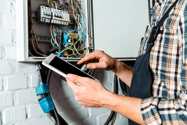 cropped view of electrician using digital tablet with blank screen near switchboard 