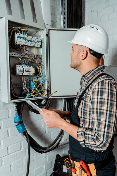 electrician in safety helmet looking at switchboard and holding digital tablet 