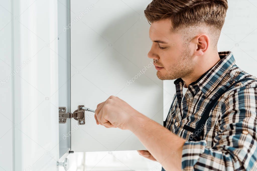 side view of handsome installer holding screwdriver near kitchen cabinet 