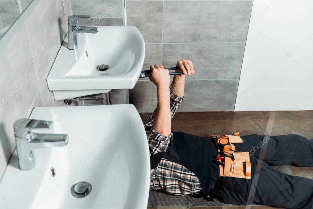 cropped view of installer holding slip joint pliers near pipe while lying on floor in bathroom 