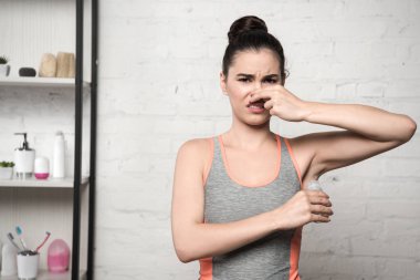 shocked woman plugging nose with hand while applying deodorant on underarm clipart