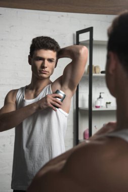 young man in white sleeveless shirt applying deodorant on underarm while looking at mirror clipart