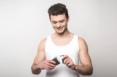 Beyaz kolsuz gömlekli gülümseyen adam gri renkte deodorant açıyor.