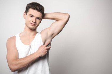 Beyaz kolsuz gömlekli yakışıklı adam koltuk altına deodorant sürüyor ve gri arka planda kameraya gülümsüyor.