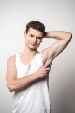 Beyaz kolsuz gömlekli yakışıklı adam koltuk altına deodorant sürüyor ve gri üzerine izole edilmiş kameraya gülümsüyor.