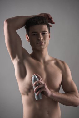 young shirtless man looking at camera while holding deodorant near underarm isolated on grey clipart
