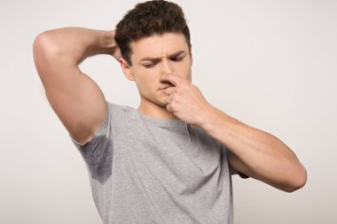 displeased man in grey t-shirt with sweaty underarm plugging nose with hand isolated on grey clipart