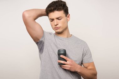 displeased man in grey t-shirt with sweaty underarm holding deodorant isolated on grey clipart