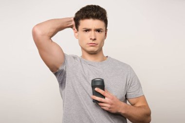 displeased man in grey t-shirt with sweaty underarm holding deodorant isolated on grey clipart