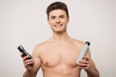 Gri renkte deodorantlar tutarken kameraya bakan tişörtsüz bir adam.