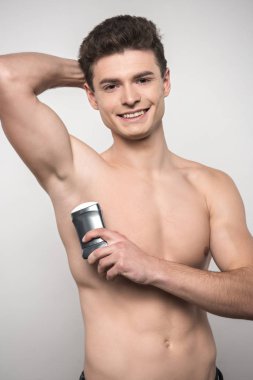 shirtless young man smiling at camera while holding deodorant near underarm isolated on grey clipart