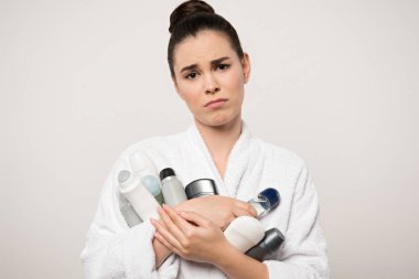 displeased woman in bathrobe holding different deodorants while looking at camera isolated on grey clipart