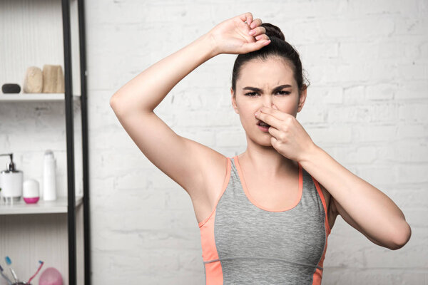 displeased woman plugging nose with hand while standing with raised hand and looking at camera