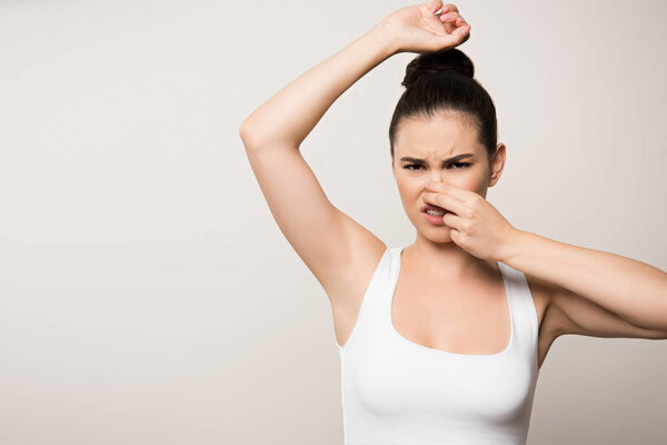 displeased woman plugging nose with hand while looking at camera isolated on grey