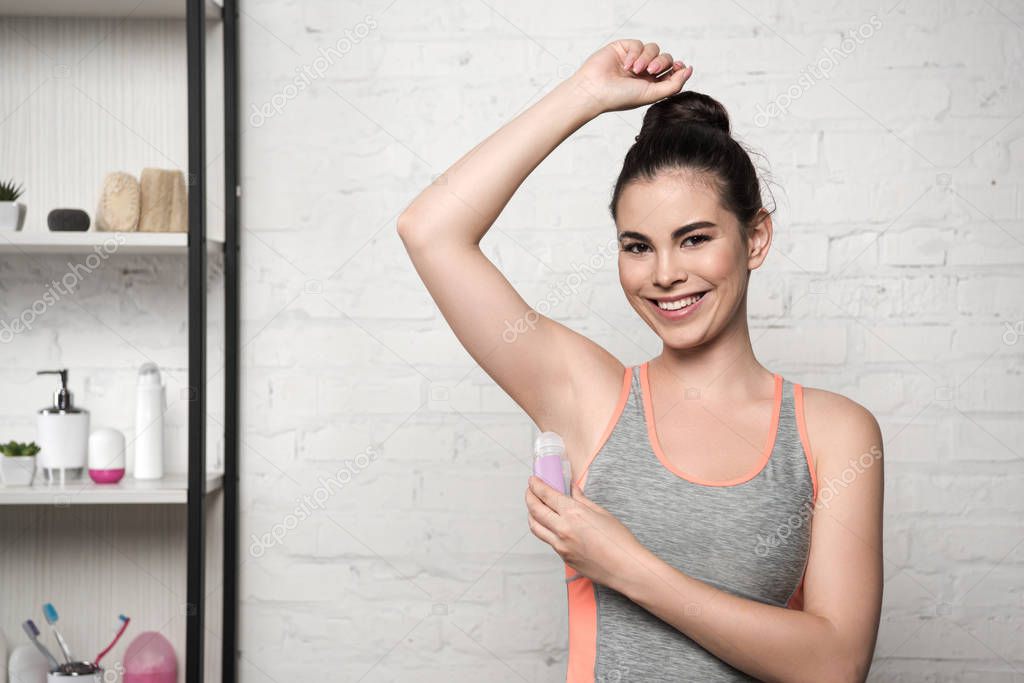happy woman smiling at camera while applying deodorant on underarm