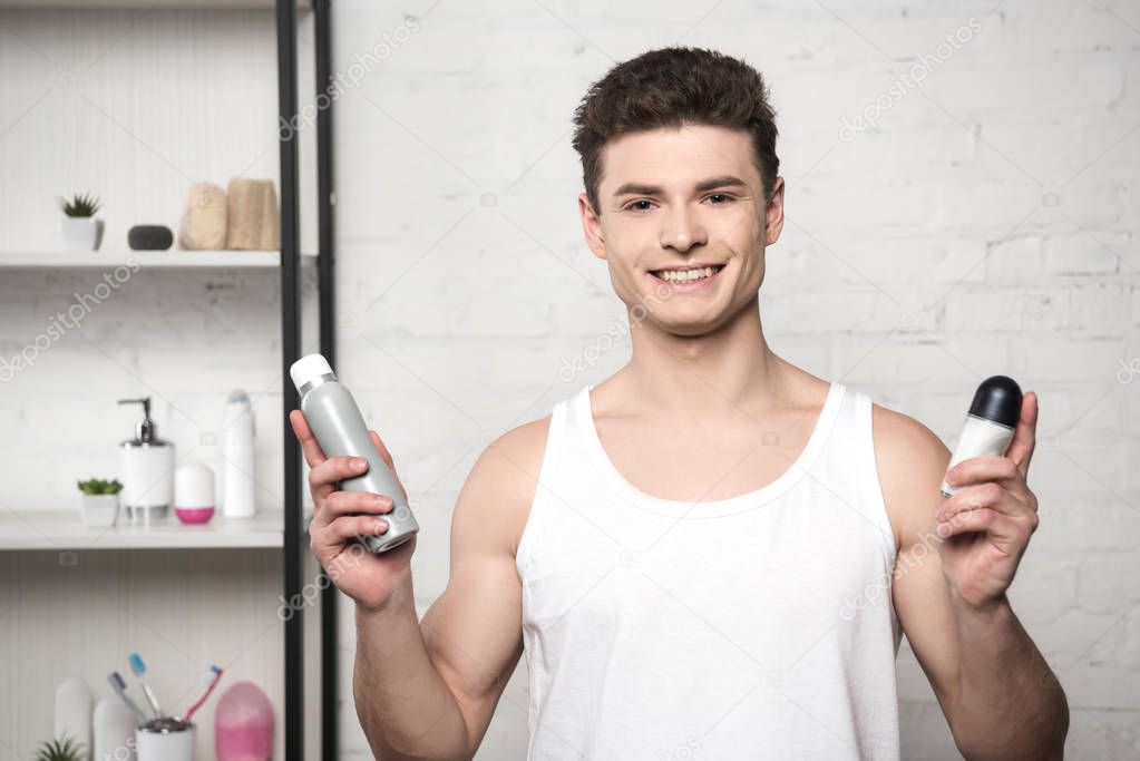 Young man in white sleeveless shirt holding deodorants and smiling at camera