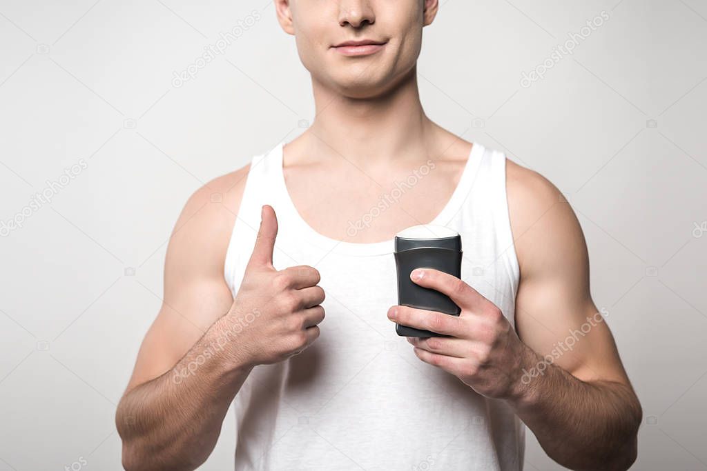 cropped view of young man in white sleeveless shirt holding deodorant and showing thumb up isolated on grey