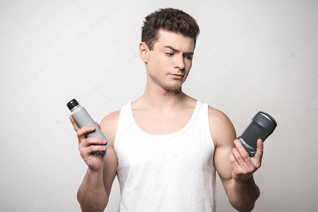 skeptical man in white sleeveless shirt holding deodorants isolated on grey
