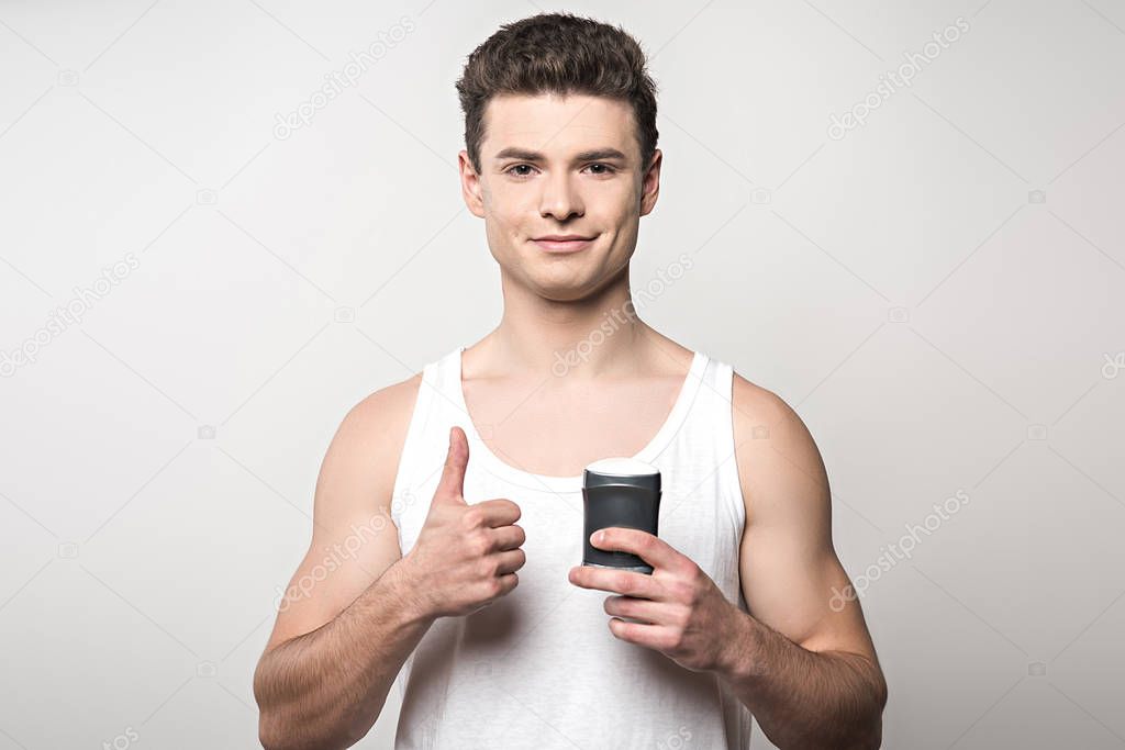 smiling man in white sleeveless shirt holding deodorant and showing thumb up isolated on grey