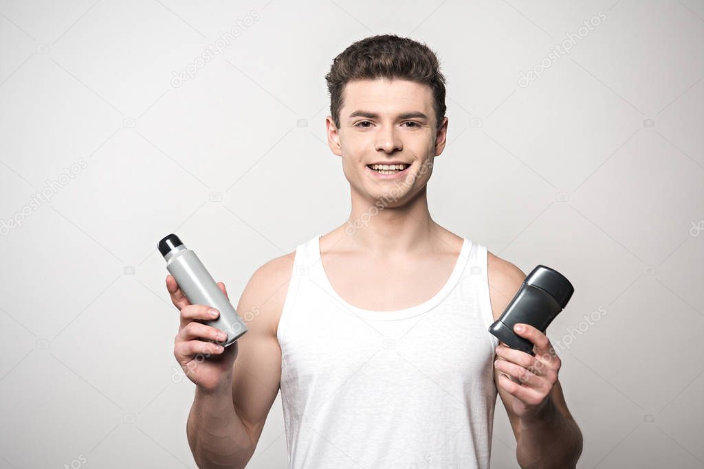 smiling man in white sleeveless shirt holding deodorants and looking at camera isolated on grey