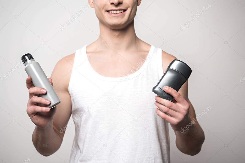 cropped view of smiling man in white sleeveless shirt holding deodorants isolated on grey