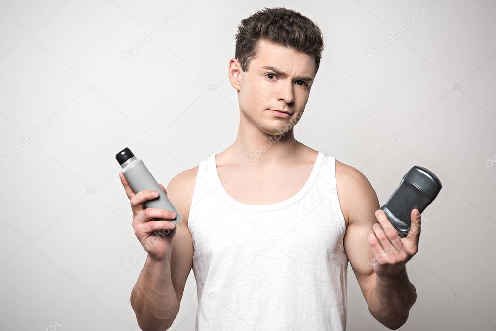 skeptical man in white sleeveless shirt holding deodorants isolated on grey