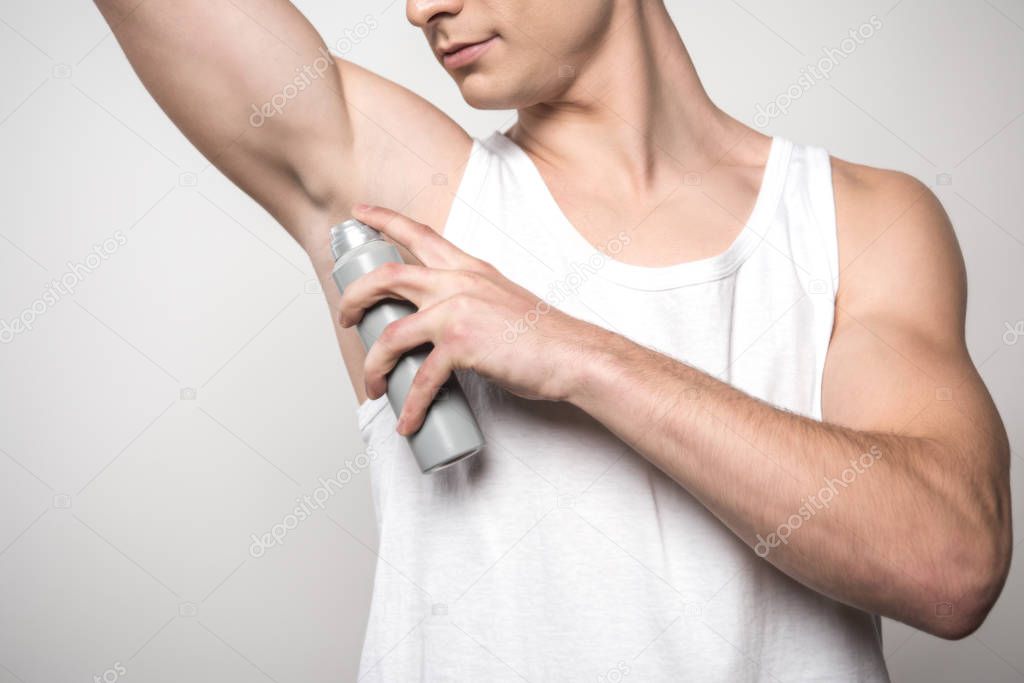 cropped view of young man in white sleeveless shirt applying deodorant on underarm isolated on grey
