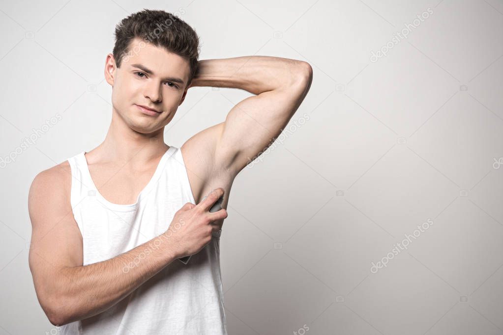 handsome man in white sleeveless shirt applying deodorant on underarm and smiling at camera on grey background