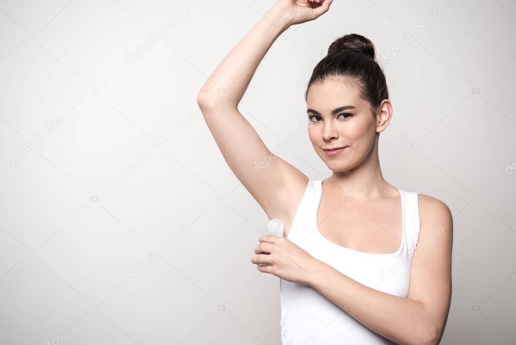 positive woman looking at camera while applying deodorant on underarm isolated on grey