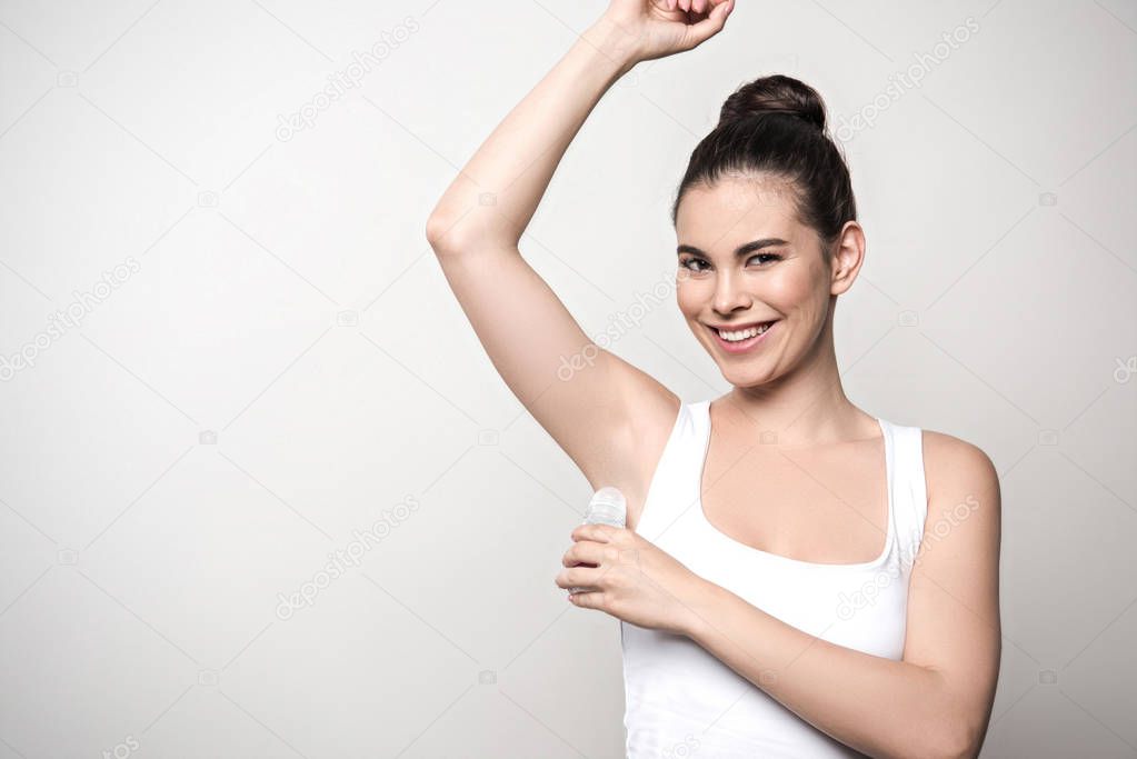 joyful woman smiling at camera while applying deodorant on underarm isolated on grey