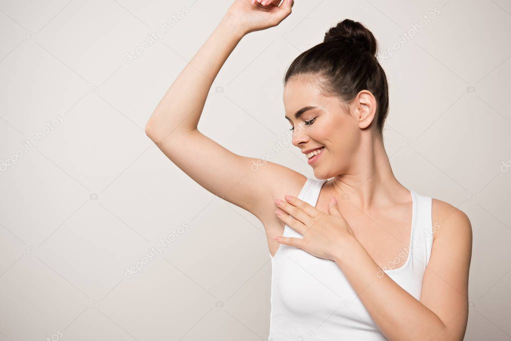 happy woman in white sleeveless shirt touching underarm isolated on grey