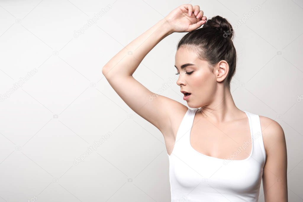 shocked woman in white sleeveless shirt looking at underarm isolated on grey