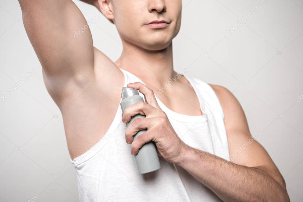 partial view of young man in white sleeveless shirt spraying deodorant on underarm isolated on grey